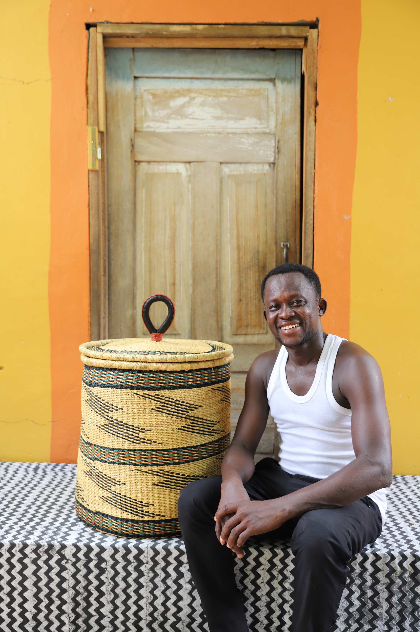 Handwoven Bolga laundry basket in natural straw with black bands, styled beside a seated man in a white vest.