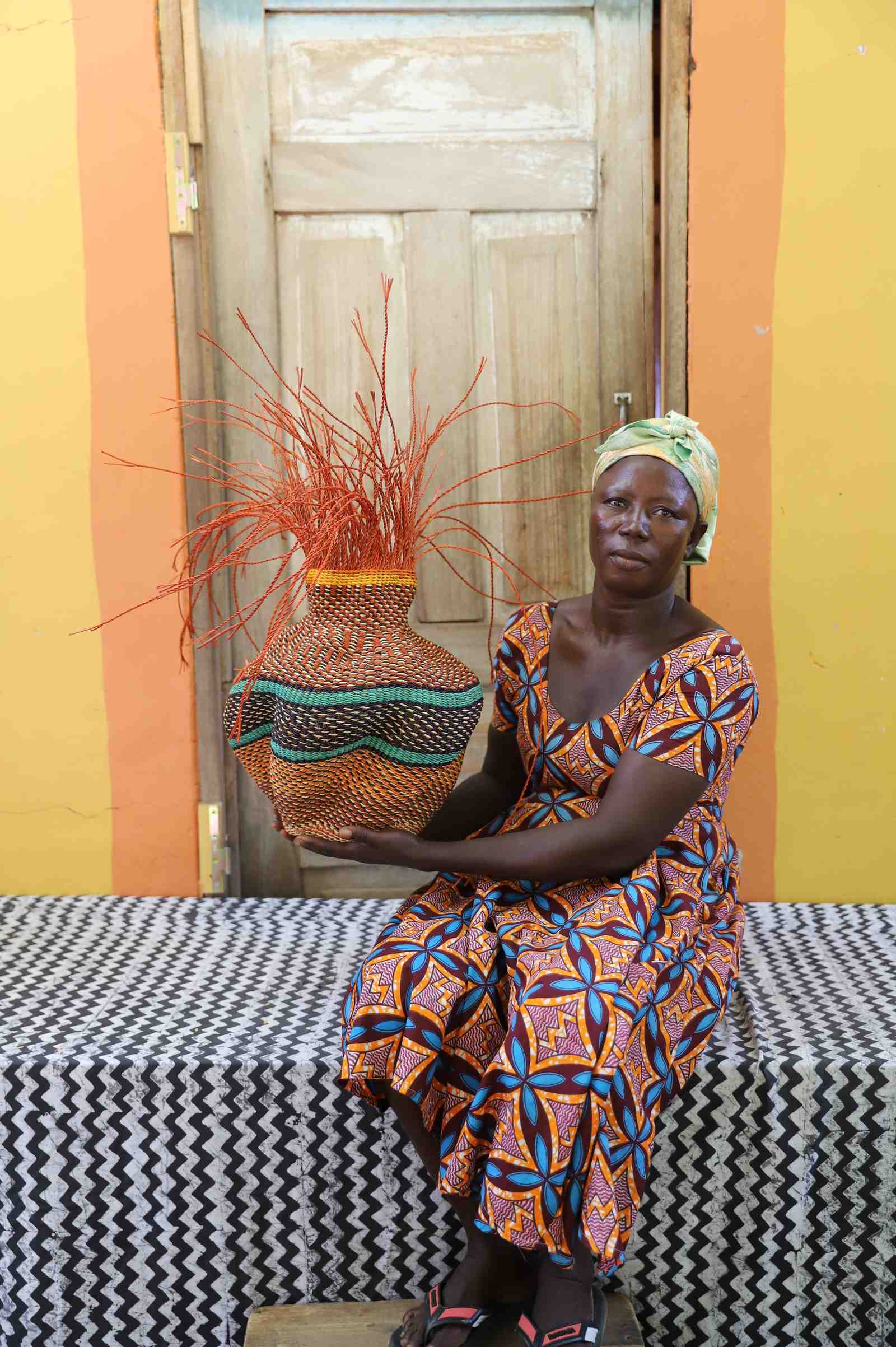 Handwoven Bolga Baobab basket in natural straw with teal scalloped bands, held by a seated woman in an orange dress.