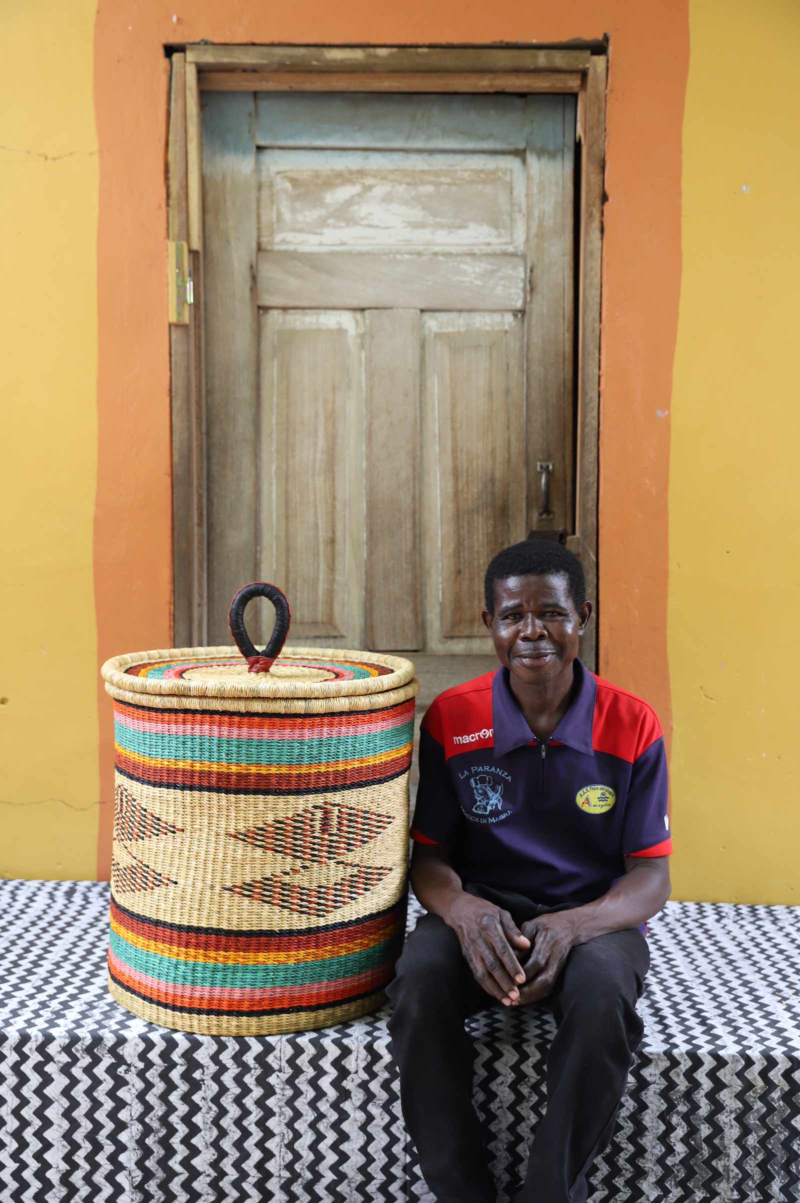 Handwoven Bolga laundry basket in multicolour bands, styled beside a seated man in red and blue.