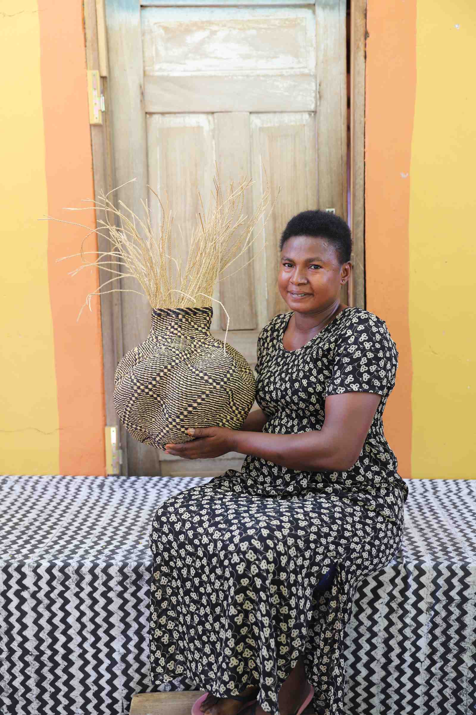 Handwoven Bolga Baobab basket in natural straw with dark diamond weave, held by a seated woman in a printed dress.