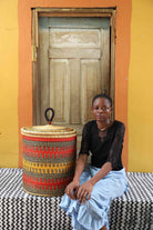 Handwoven Bolga laundry basket in deep red with orange and yellow bands, styled beside a seated woman.
