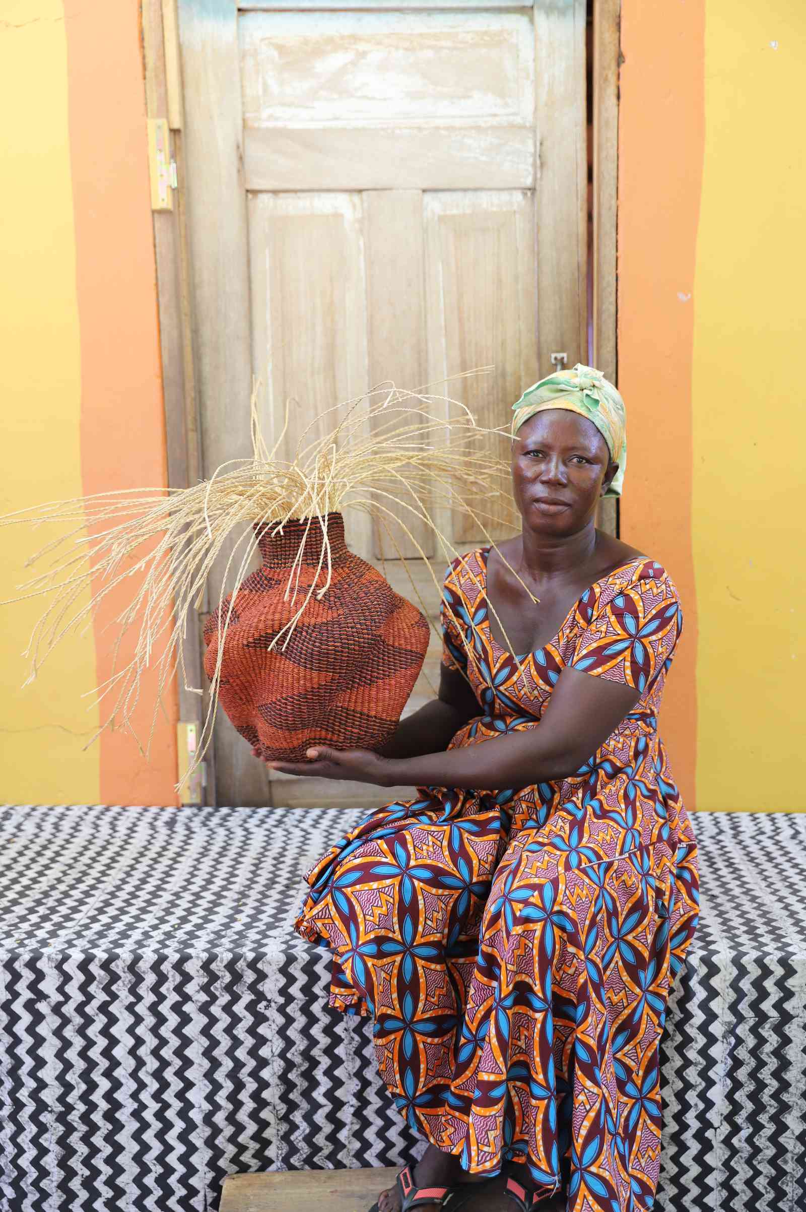 Handwoven Bolga Baobab basket in rust-red with cream grass sprigs, held by a seated woman in a printed dress.