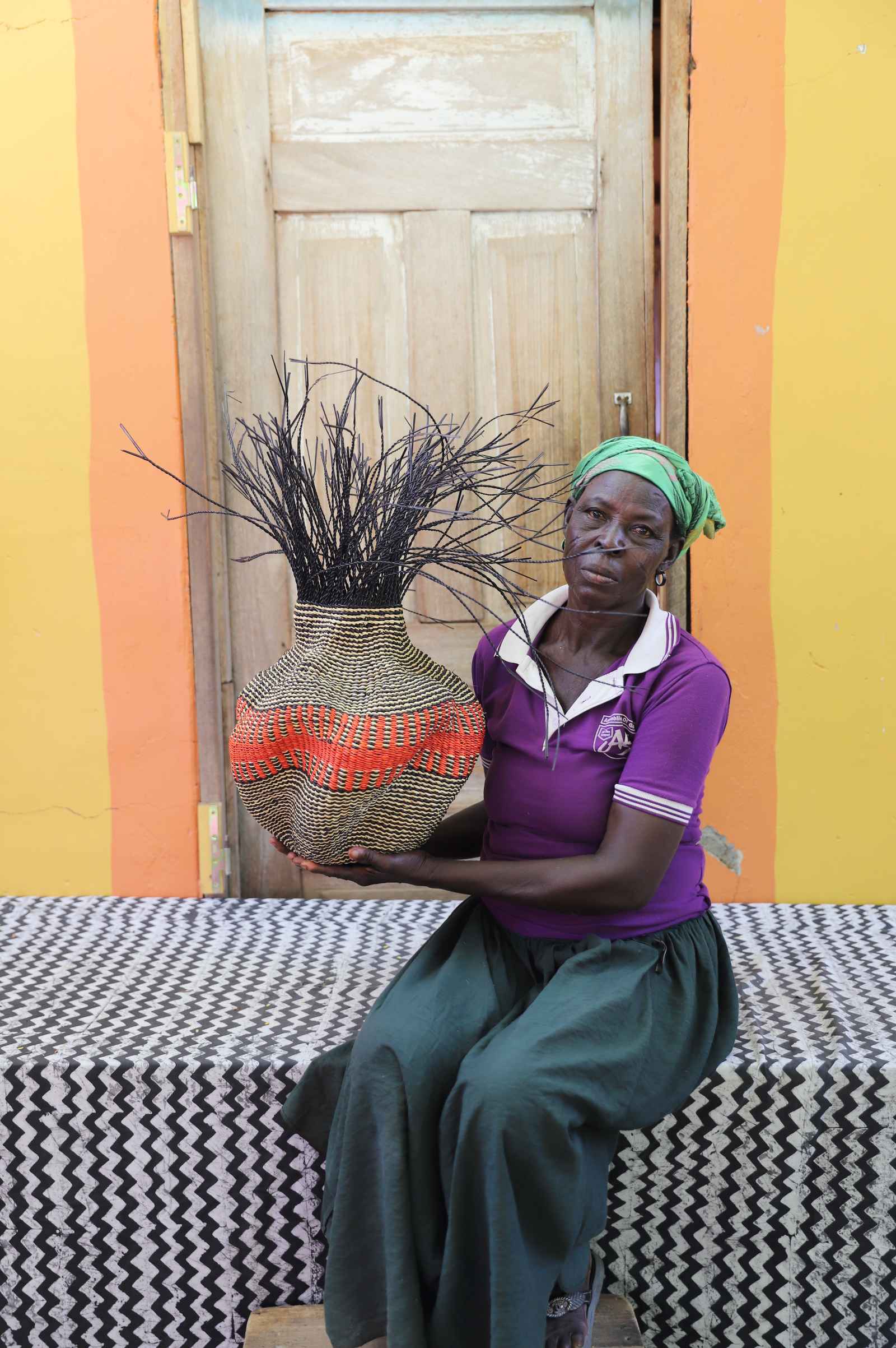 Handwoven Bolga Baobab basket in natural straw with a red central band, held by a seated woman in purple.