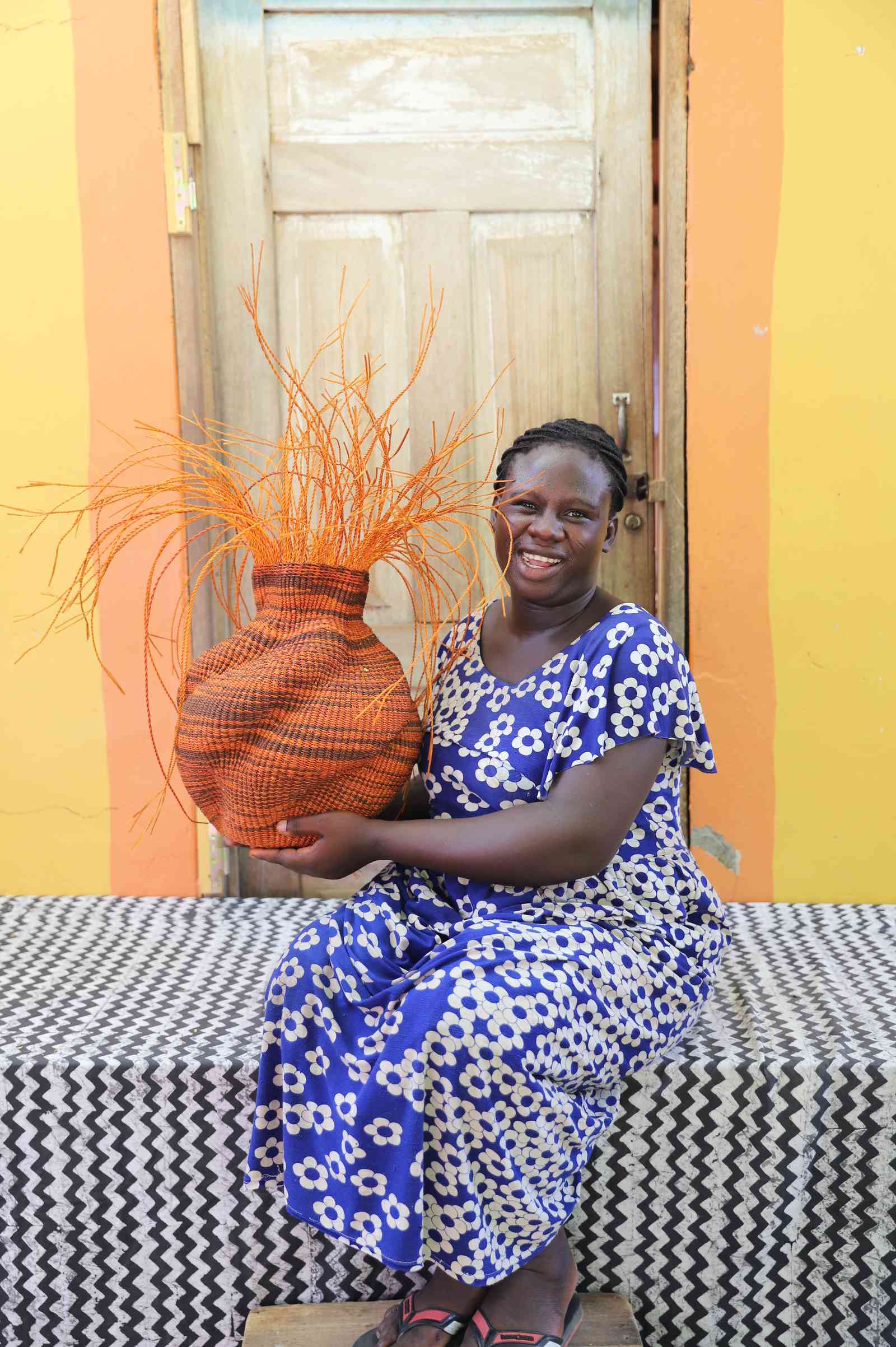 Handwoven Bolga Baobab basket in dyed orange with orange grass sprigs, held by a seated woman in blue floral.