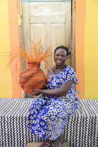 Handwoven Bolga Baobab basket in dyed orange with orange grass sprigs, held by a seated woman in blue floral.