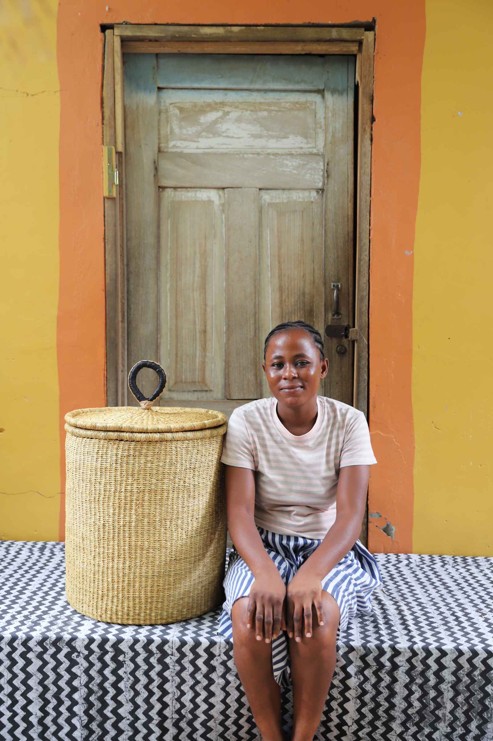 Handwoven Bolga laundry basket with coiled rim and lid in natural elephant grass, styled beside a seated woman.