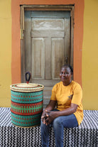 Handwoven Bolga laundry basket in teal, red and green bands, styled beside a seated woman in yellow.