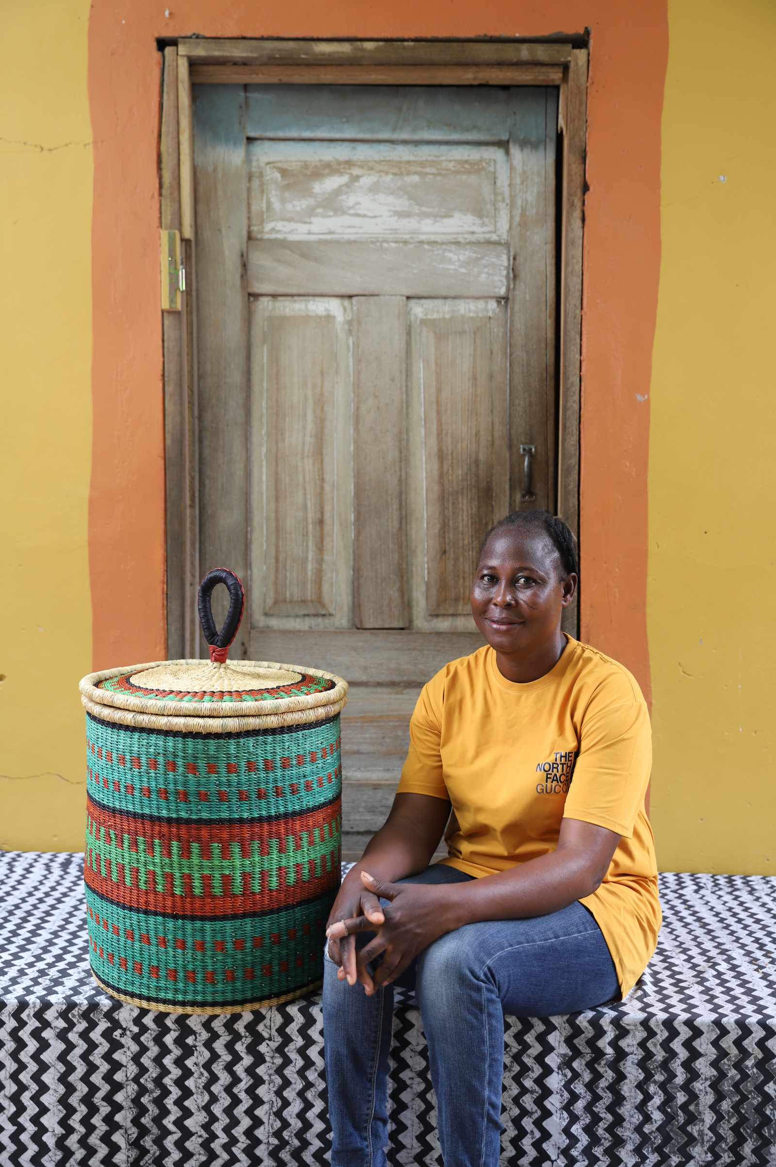 Handwoven Bolga laundry basket in teal, red and green bands, styled beside a seated woman in yellow.