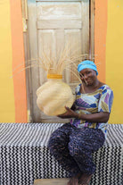 Handwoven Bolga Baobab basket in natural straw with grass sprigs on top, held by a seated woman in printed dress.