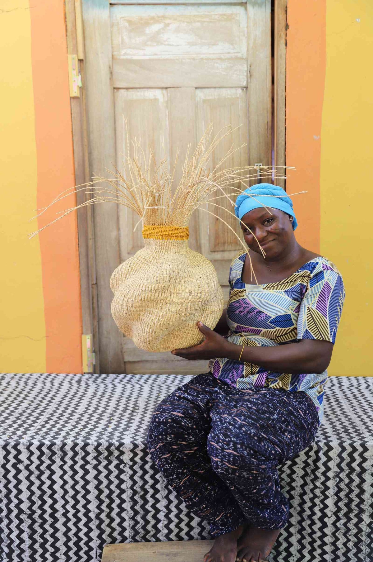 Handwoven Bolga Baobab basket in natural straw with grass sprigs on top, held by a seated woman in printed dress.