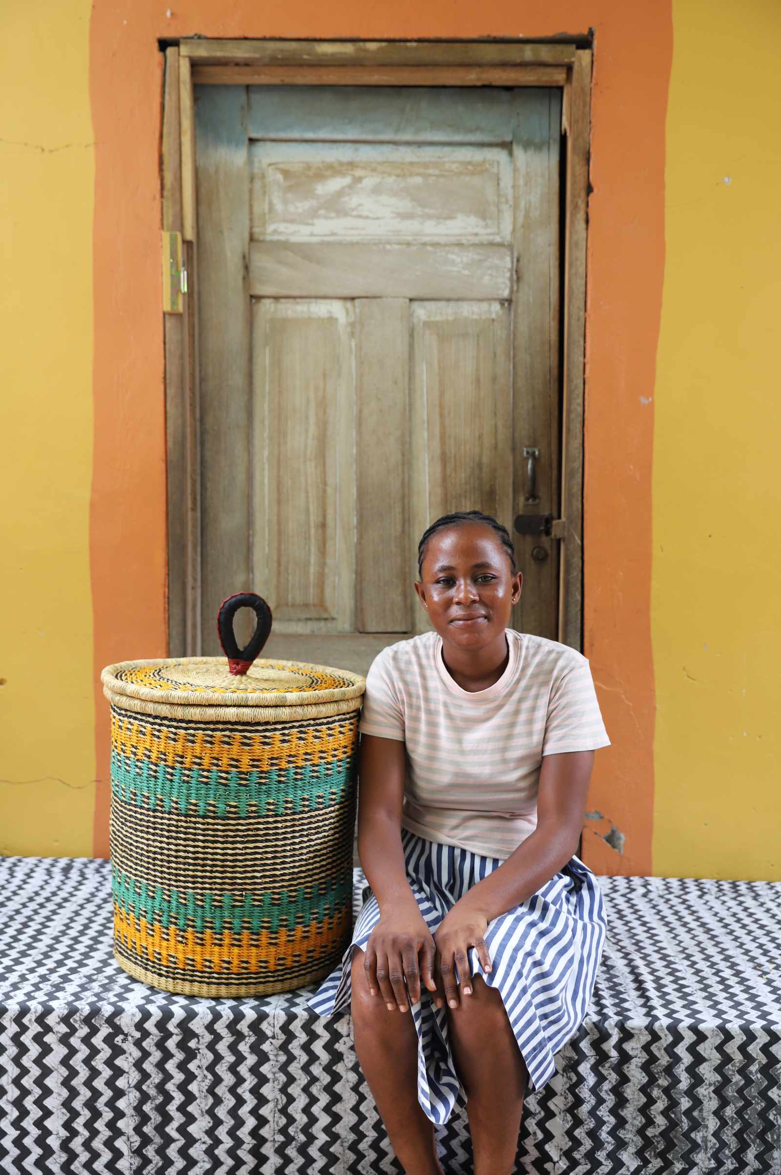 Handwoven Bolga laundry basket in natural straw with orange and teal bands, styled beside a seated woman.