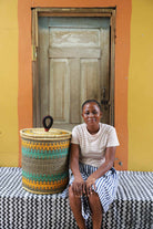 Handwoven Bolga laundry basket in natural straw with orange and teal bands, styled beside a seated woman.