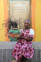 Handwoven Bolga Baobab basket in natural straw with teal bands, held by a seated woman in a red printed dress.