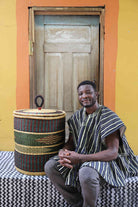Handwoven Bolga laundry basket in natural straw with a dark green band, styled beside a seated man in stripes.