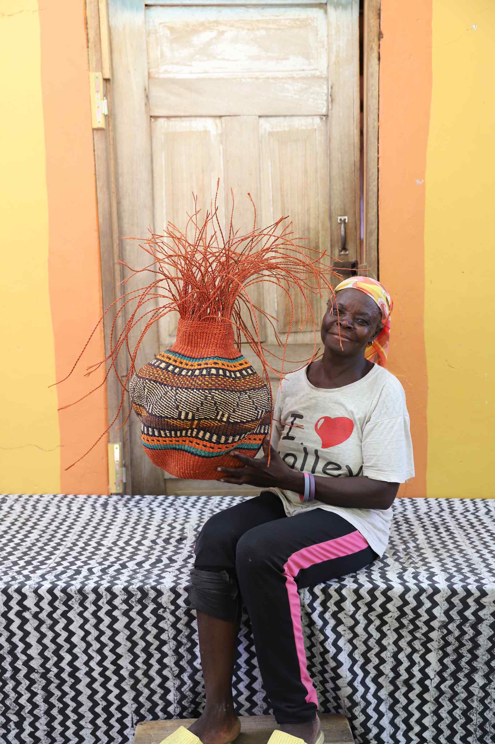 Handwoven Bolga Baobab basket in rust-orange with multicolour band, held by a seated woman in a striped top.