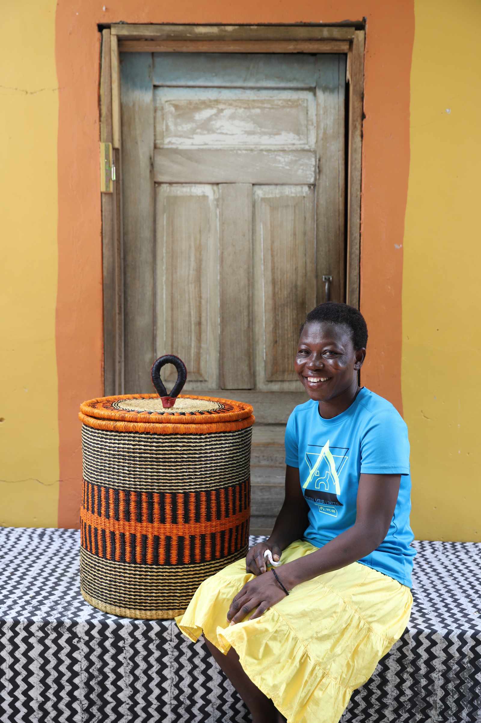 Handwoven Bolga laundry basket in natural straw with an orange band, styled beside a seated woman in yellow.
