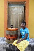 Handwoven Bolga laundry basket in natural straw with an orange band, styled beside a seated woman in yellow.