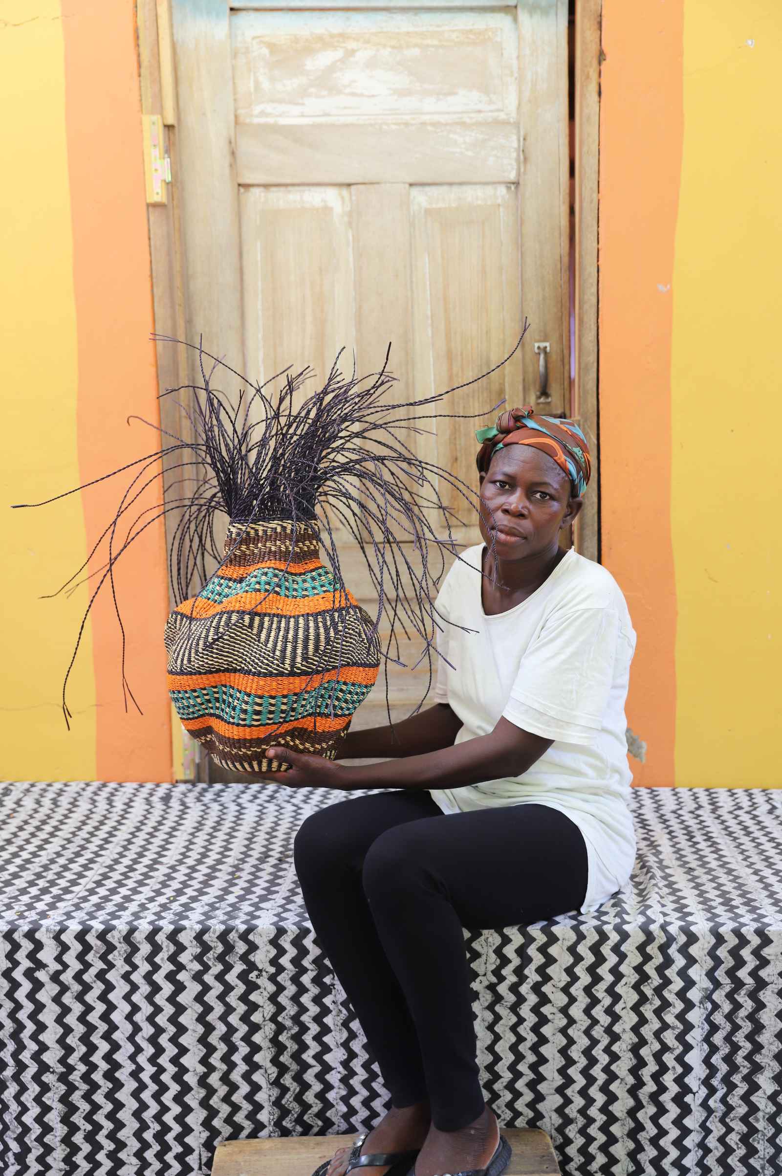 Handwoven Bolga Baobab basket in natural straw with orange and teal bands, held by a seated woman in white.