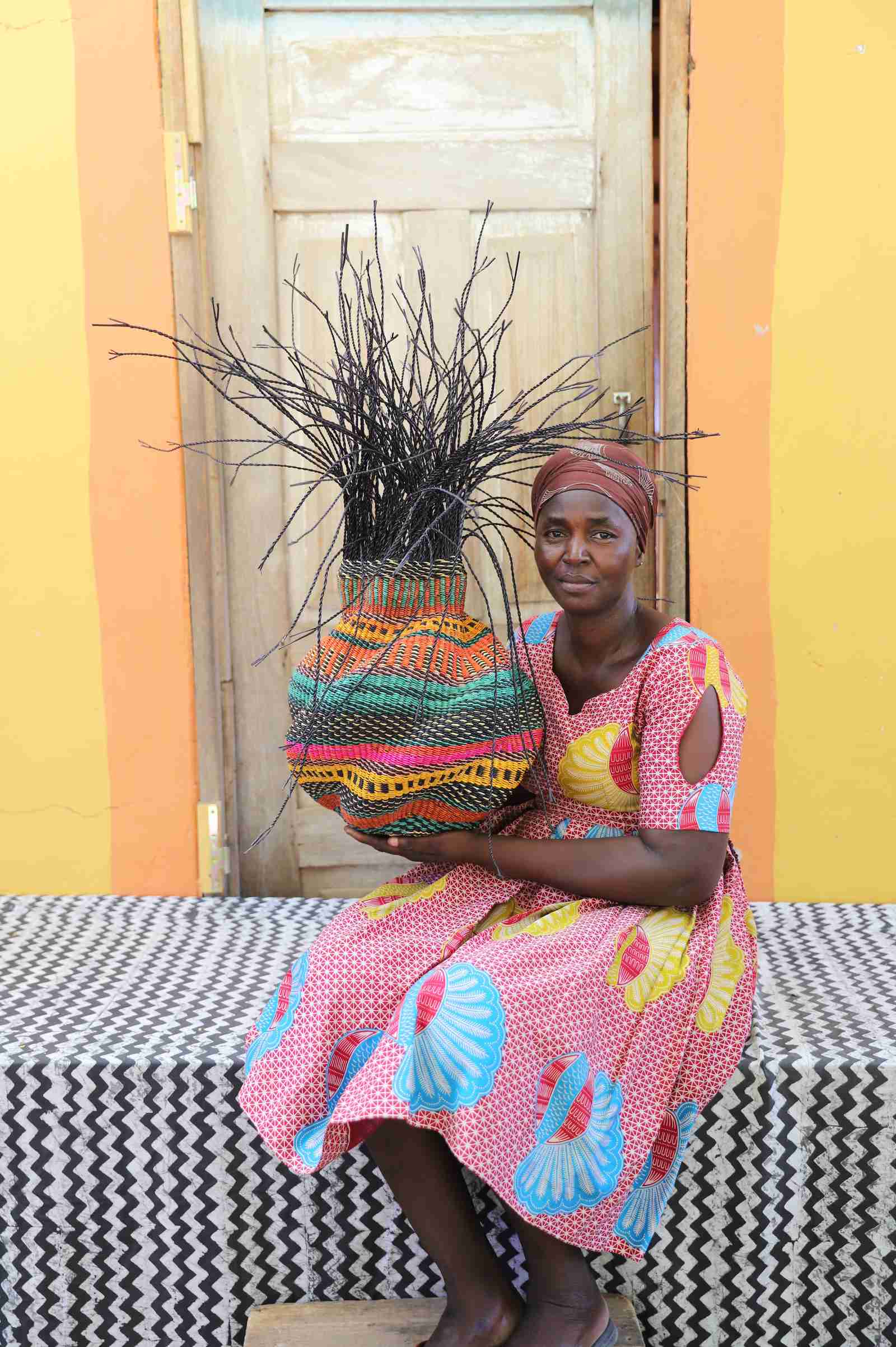 Handwoven Bolga Baobab basket with pink, teal and yellow bands, held by a seated woman in pink.