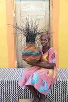 Handwoven Bolga Baobab basket with pink, teal and yellow bands, held by a seated woman in pink.