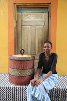 Handwoven Bolga laundry basket with lid in deep red with a black accent band, styled beside a seated woman.