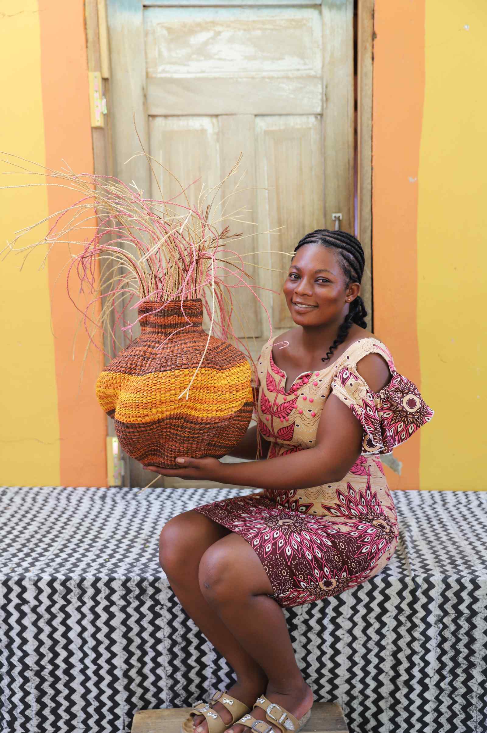 Handwoven Bolga Baobab basket with dark brown base and orange bands, held by a seated woman in a printed dress.