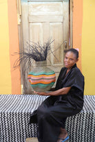 Handwoven Bolga Baobab basket with orange and teal wave stripes and dark sprigs, held by a seated woman in black.