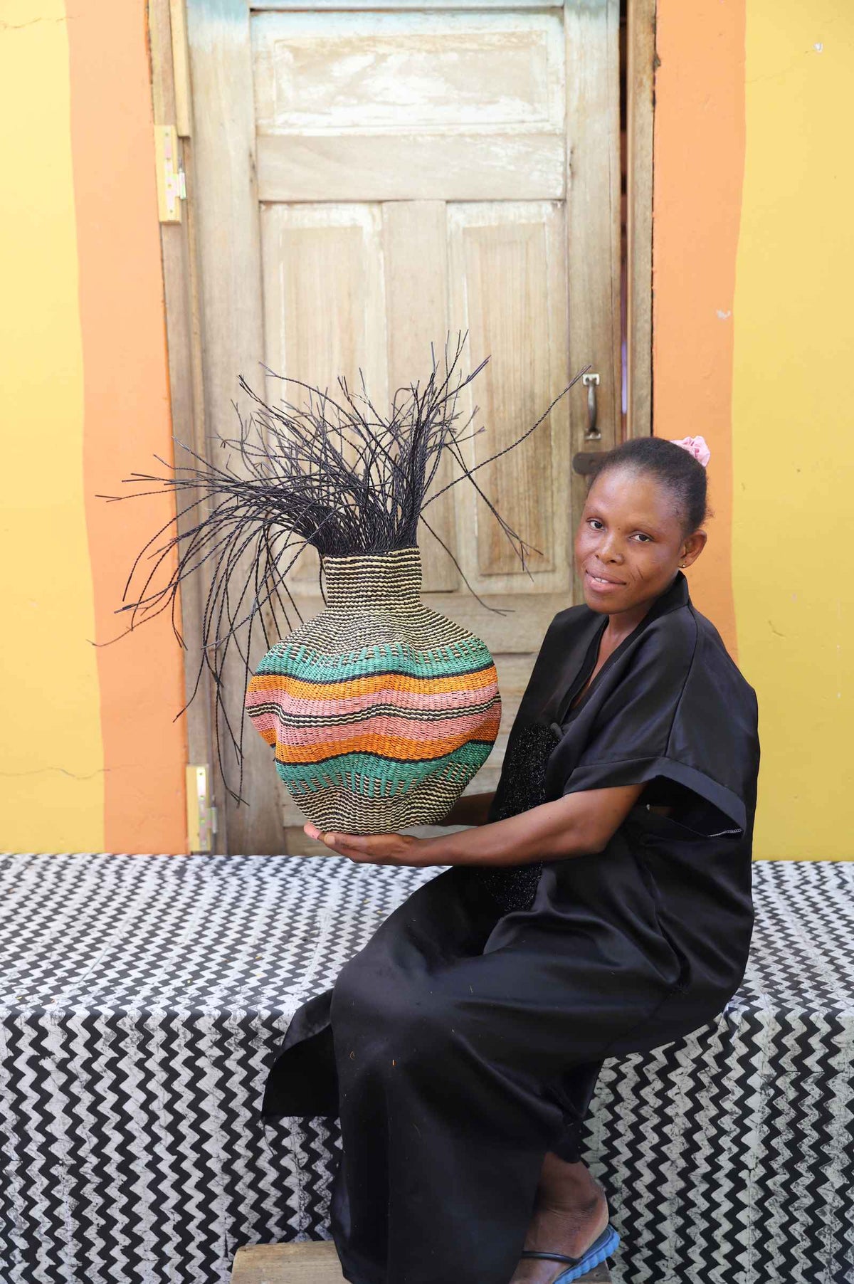 Handwoven Bolga Baobab basket with orange and teal wave stripes and dark sprigs, held by a seated woman in black.