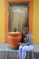 Handwoven Bolga laundry basket with lid in orange and teal, styled beside a seated woman.