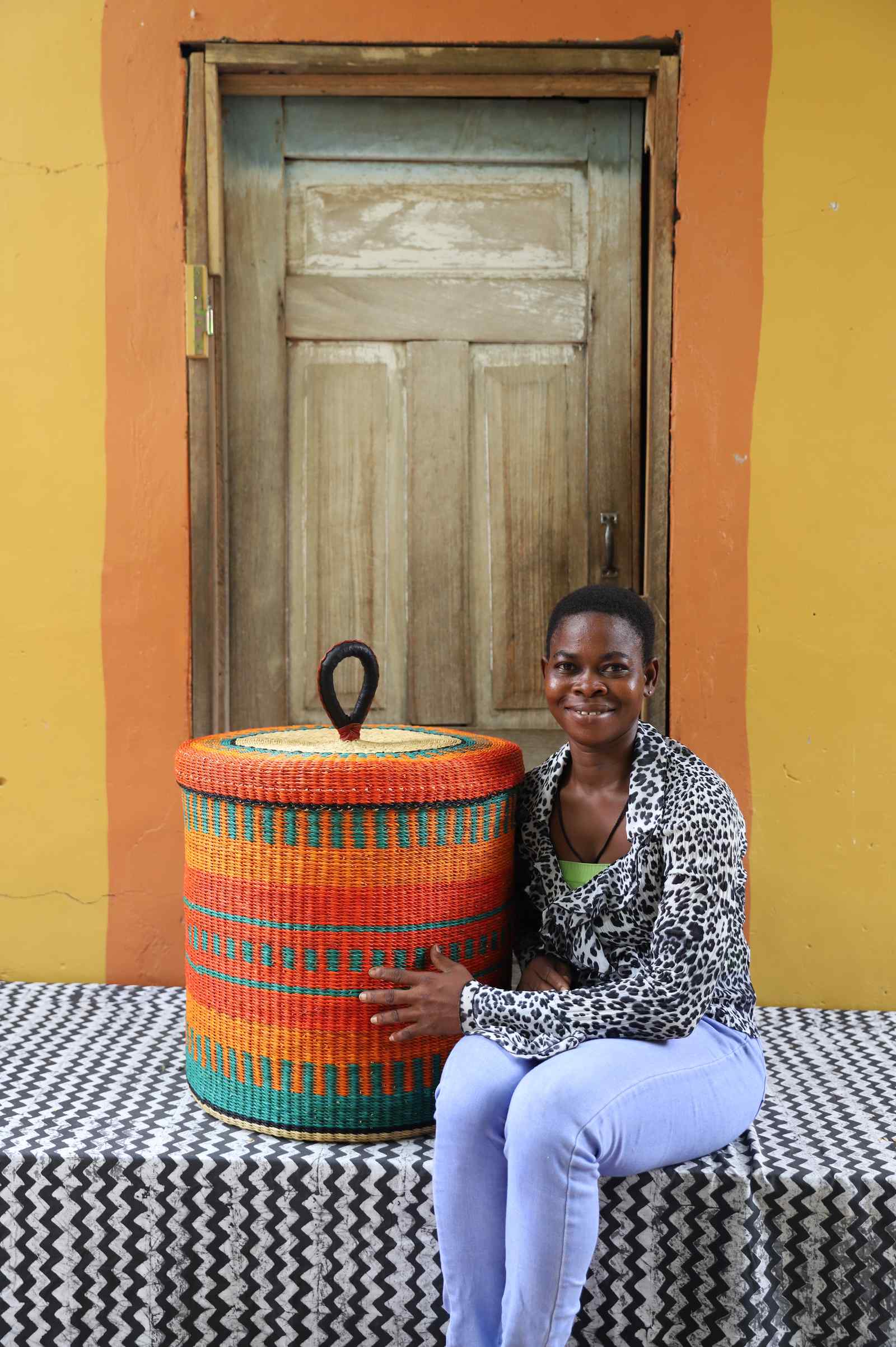 Handwoven Bolga laundry basket with lid in orange and teal, styled beside a seated woman.