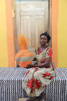 Handwoven Bolga Baobab basket in dyed orange with orange grass sprigs, held by a seated woman in a floral dress.