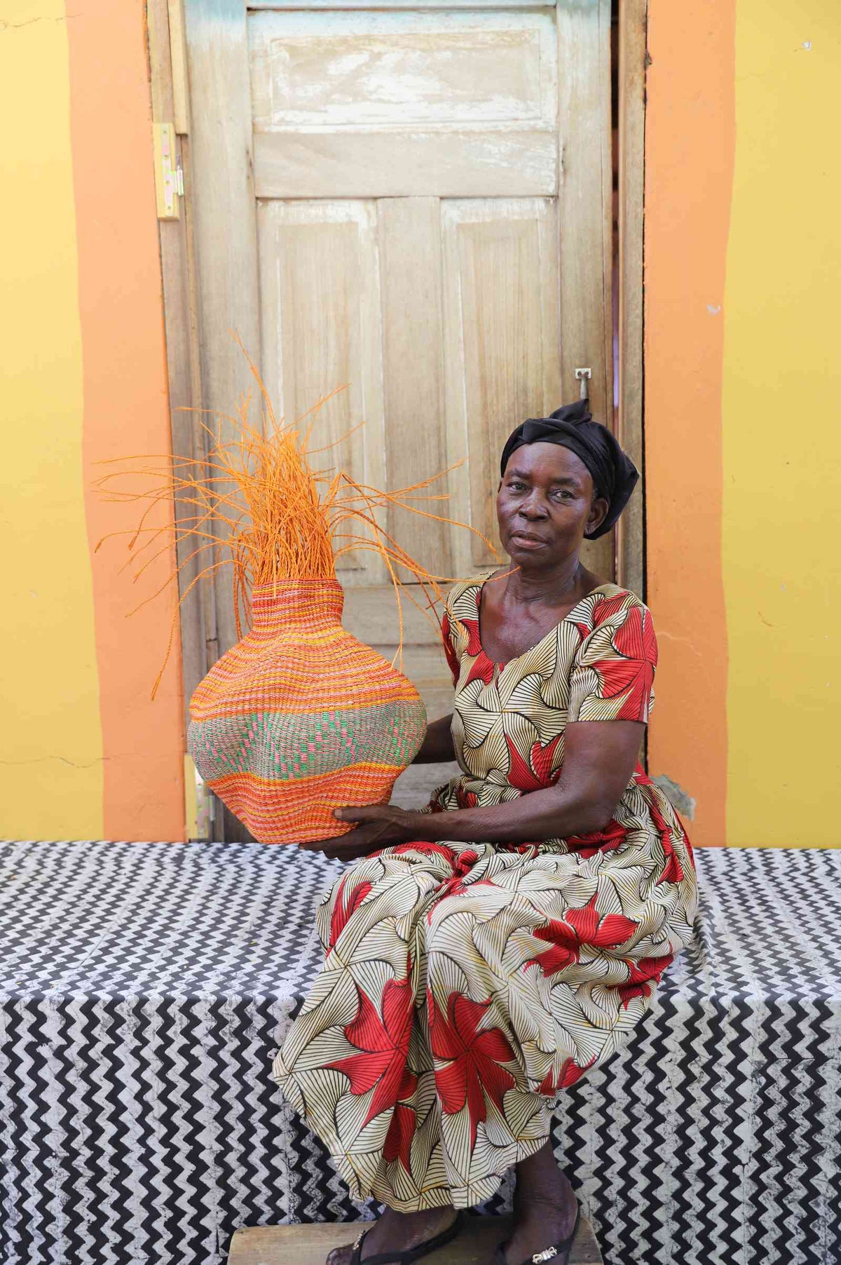 Handwoven Bolga Baobab basket in dyed orange with orange grass sprigs, held by a seated woman in a floral dress.