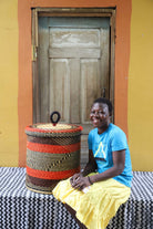 Handwoven Bolga laundry basket with lid in red and black bands, styled beside a seated woman in yellow.