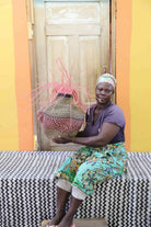 Handwoven Bolga Baobab basket in natural straw with rust-red band and pink sprigs, held by a seated woman.