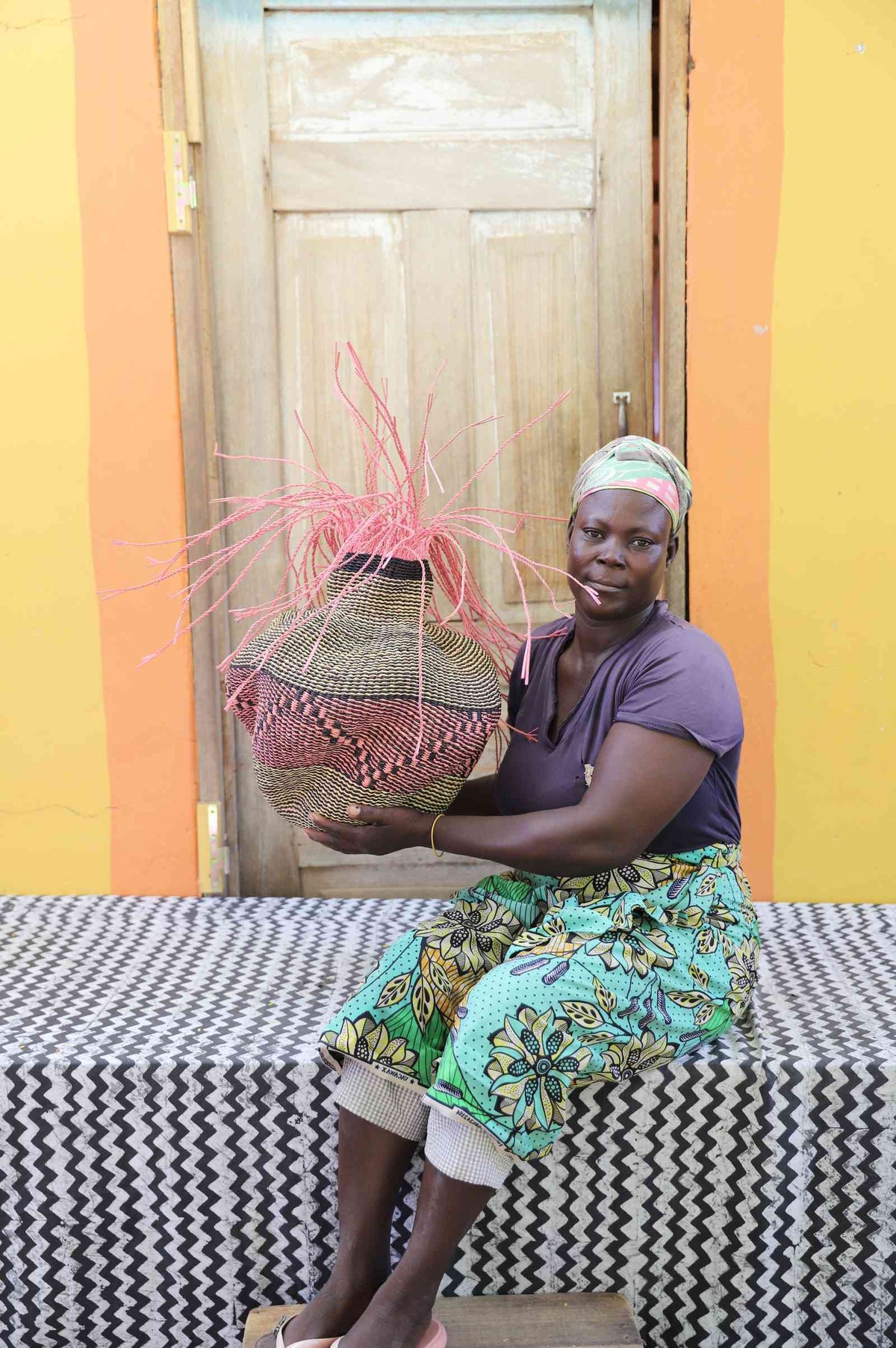 Handwoven Bolga Baobab basket in natural straw with rust-red band and pink sprigs, held by a seated woman.