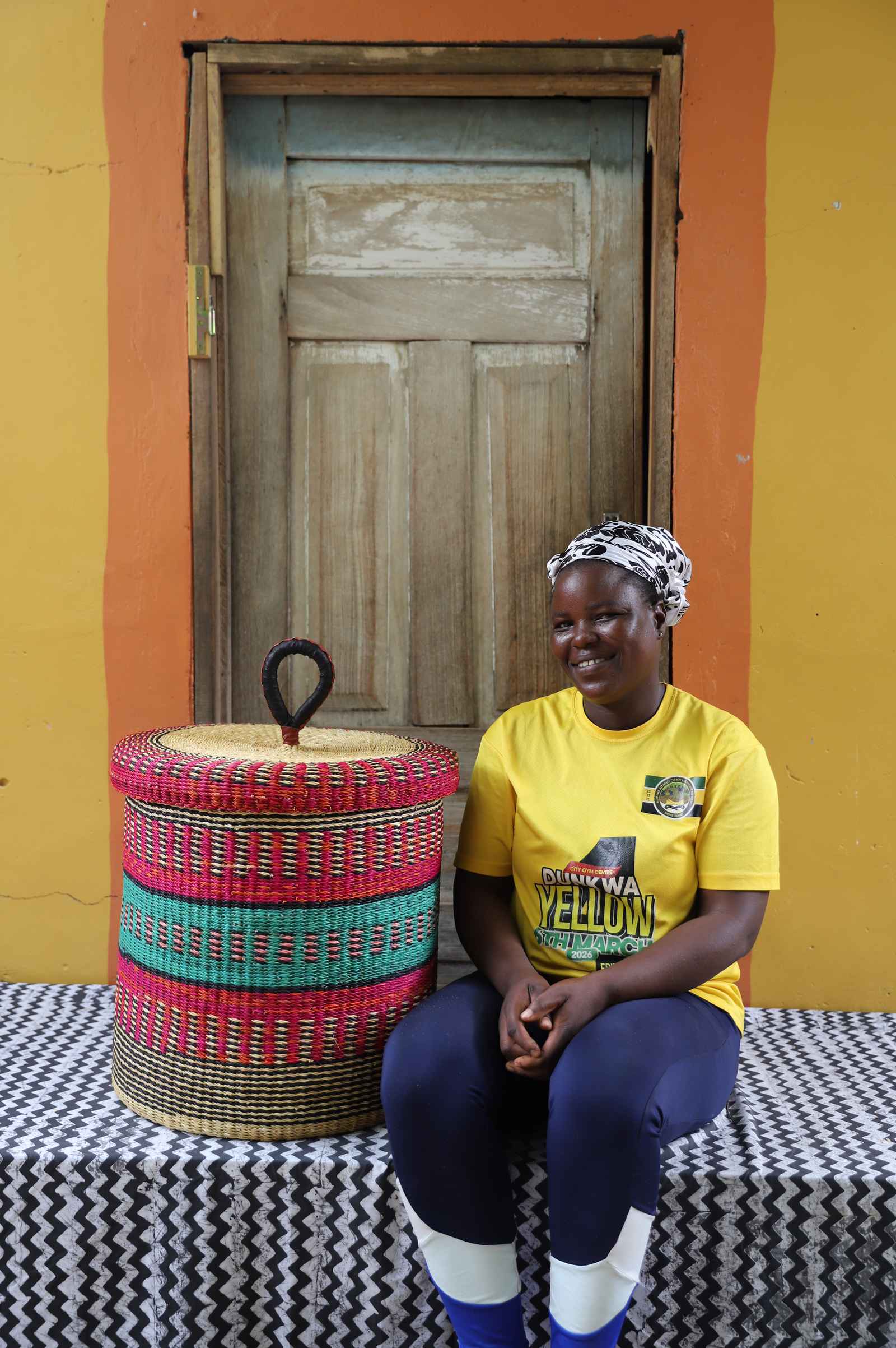 Handwoven Bolga laundry basket with lid in red and green bands, styled beside a seated woman in a yellow top.