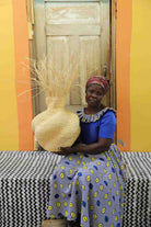 Handwoven Bolga Baobab basket in natural straw with grass sprigs, held by a seated woman in blue.