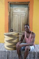 Handwoven Bolga laundry basket with lid in natural straw with a black zigzag pattern, styled beside a seated man.