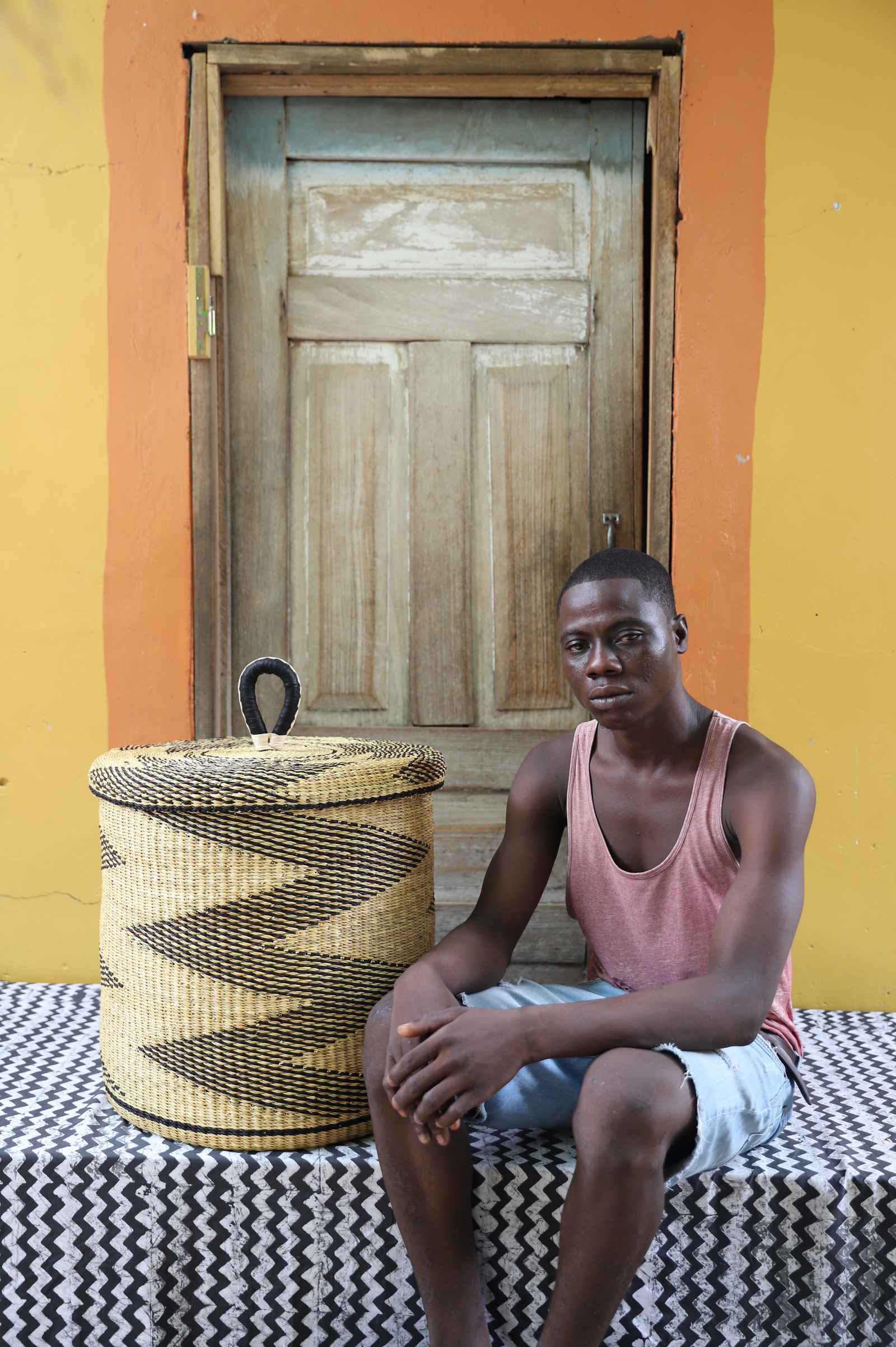 Handwoven Bolga laundry basket with lid in natural straw with a black zigzag pattern, styled beside a seated man.