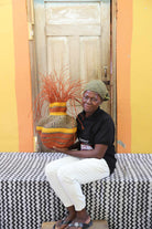 Handwoven Bolga Baobab basket in natural straw with orange band and grass sprigs, held by a seated woman.