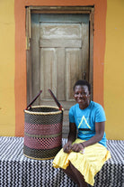 Handwoven open-top Bolga laundry basket in natural straw with black and red bands, styled beside a seated woman.