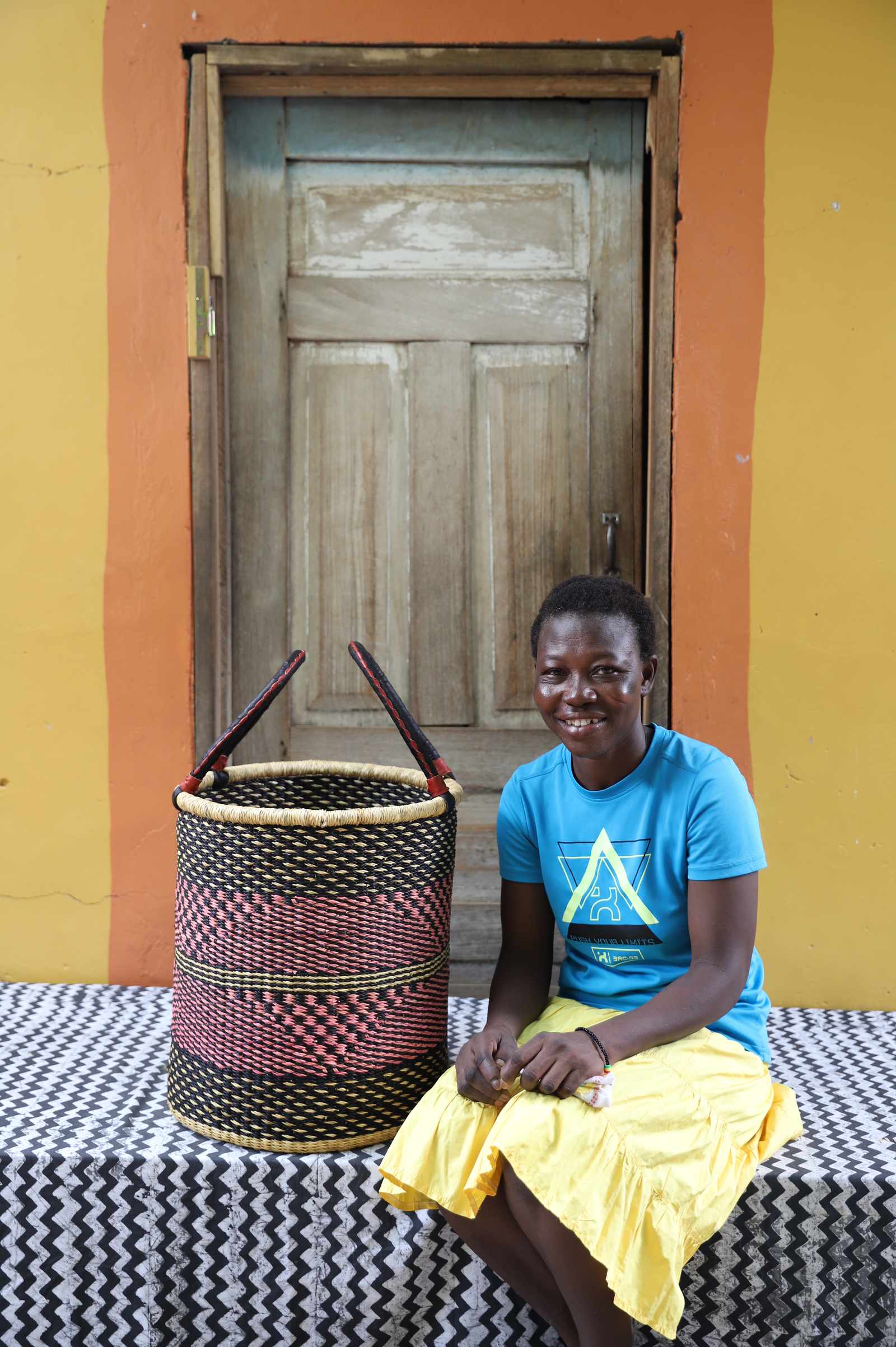 Handwoven open-top Bolga laundry basket in natural straw with black and red bands, styled beside a seated woman.