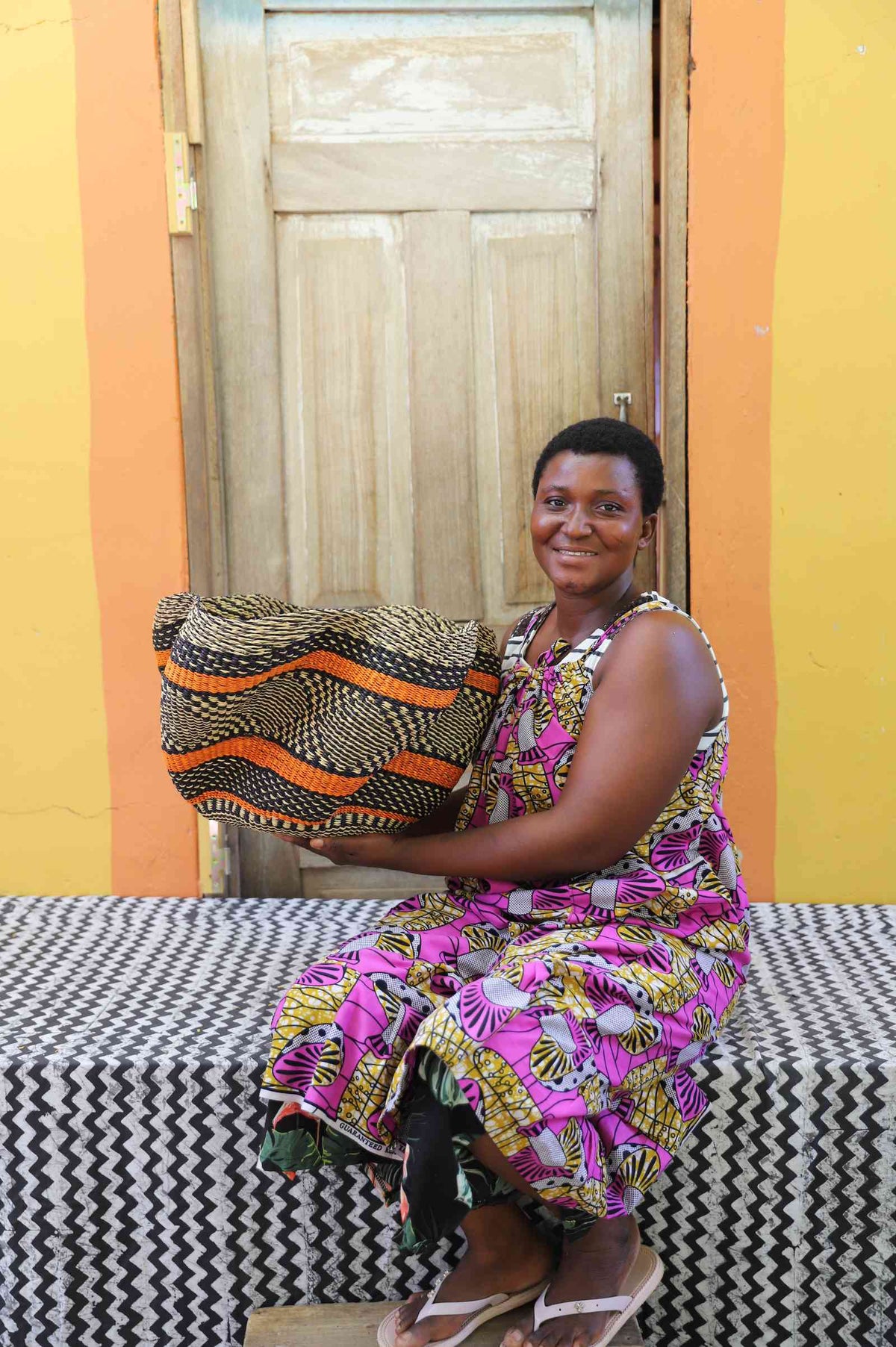 Handwoven Bolga wave basket in orange and dark wave stripes, held by a seated woman in a printed dress.