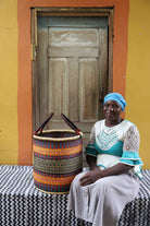 Handwoven open-top Bolga laundry basket in natural straw with orange, yellow and black stripes, styled beside a seated woman.