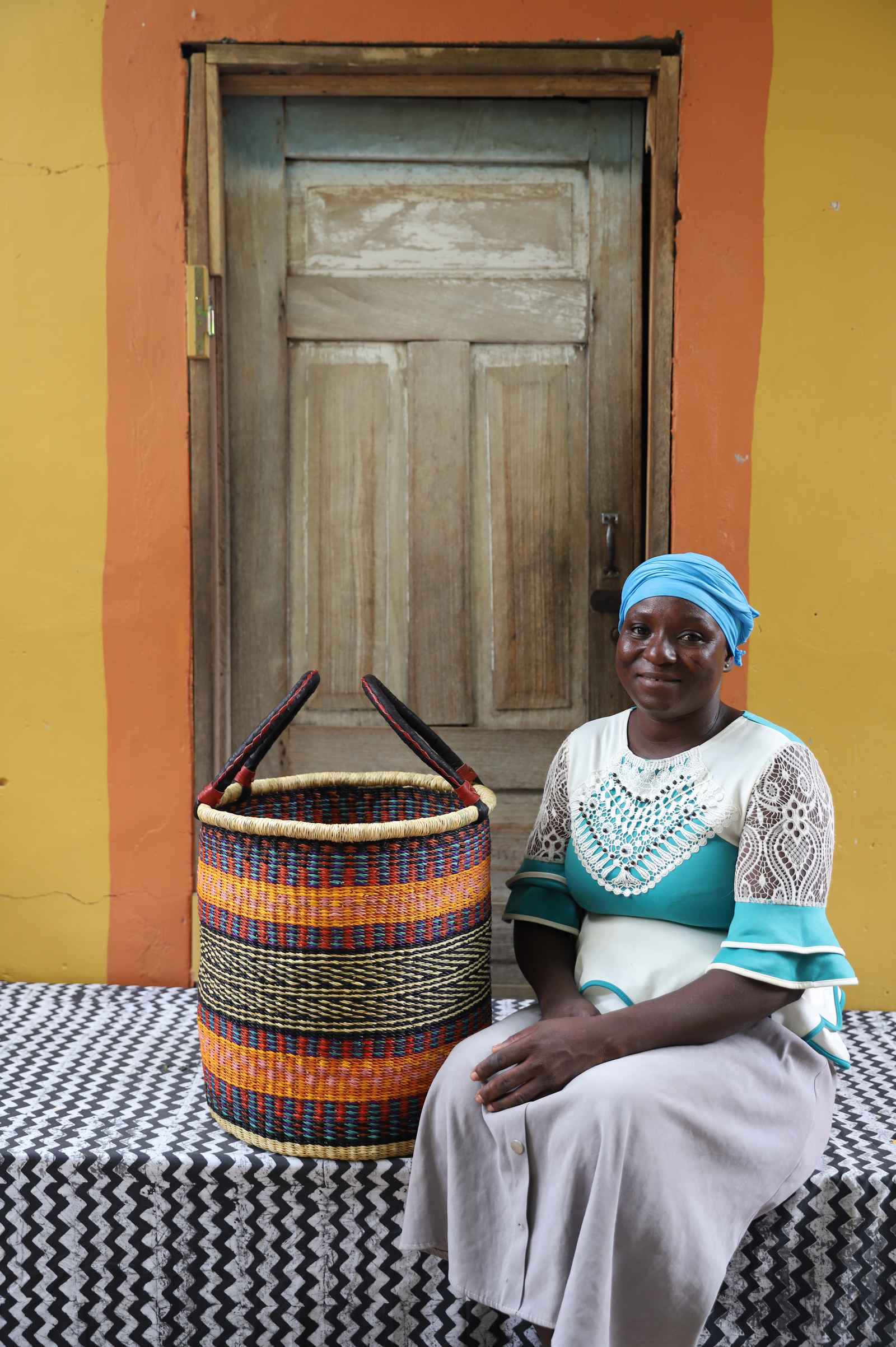 Handwoven open-top Bolga laundry basket in natural straw with orange, yellow and black stripes, styled beside a seated woman.