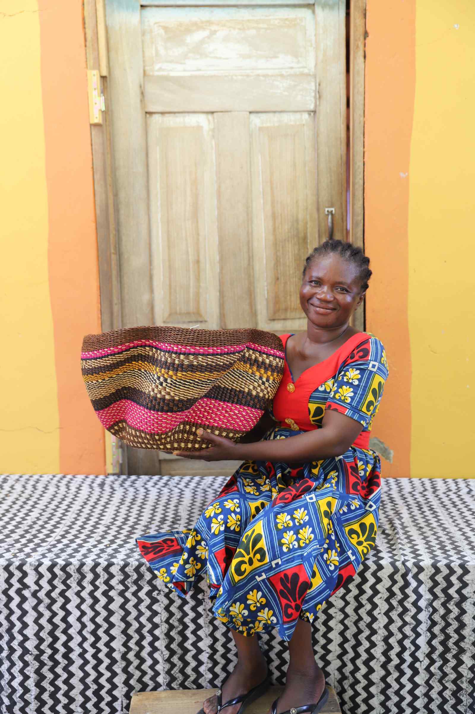 Handwoven Bolga wave basket in pink and black wavy stripes, held by a seated woman in a printed dress.