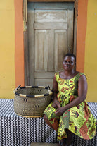 Handwoven Bolga storage basket in natural straw with a dark motif and banded rim, styled beside a seated woman.