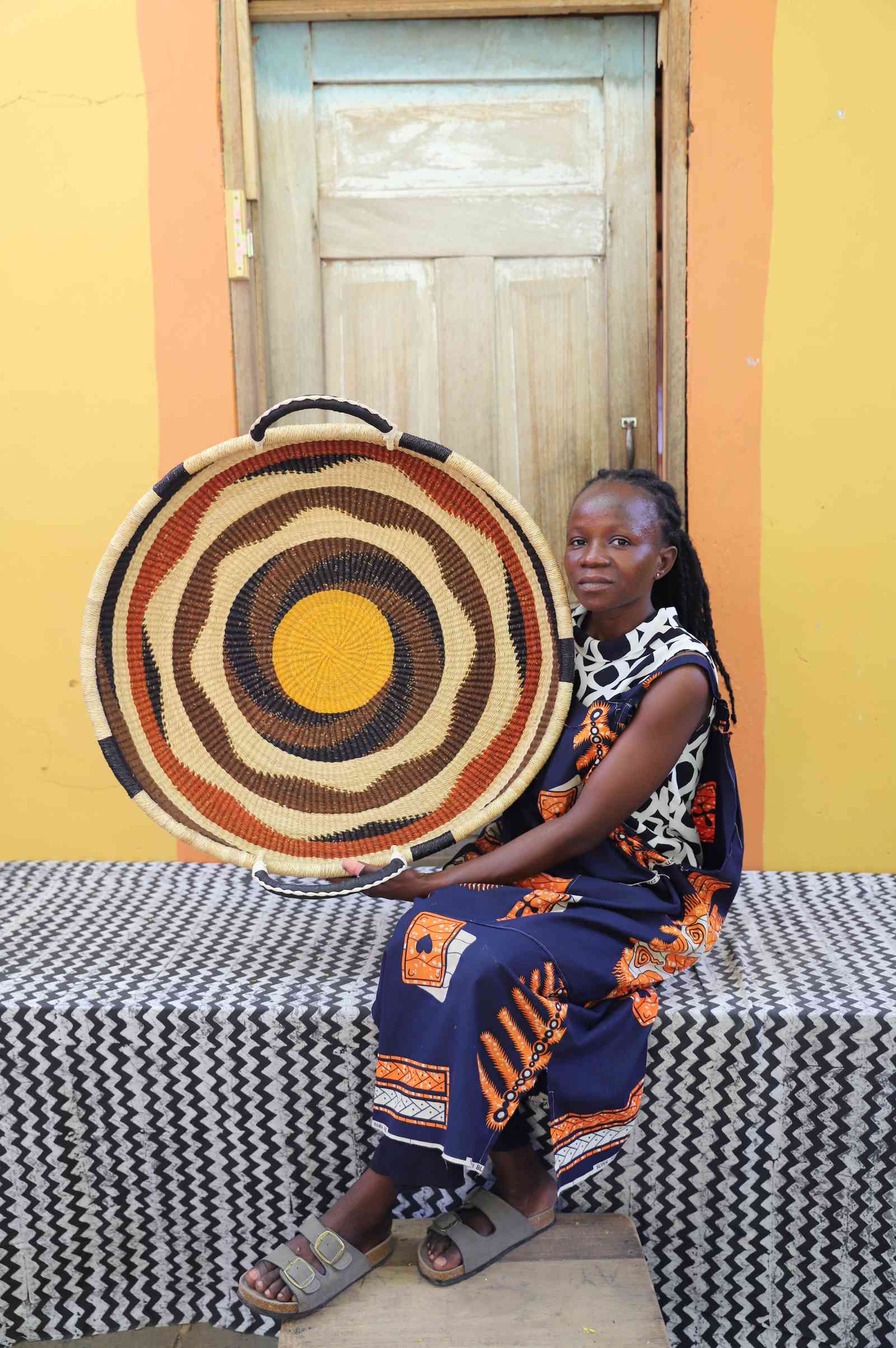 Large handwoven Bolga wall tray disc with brown and orange bands and yellow centre, held by a seated woman.