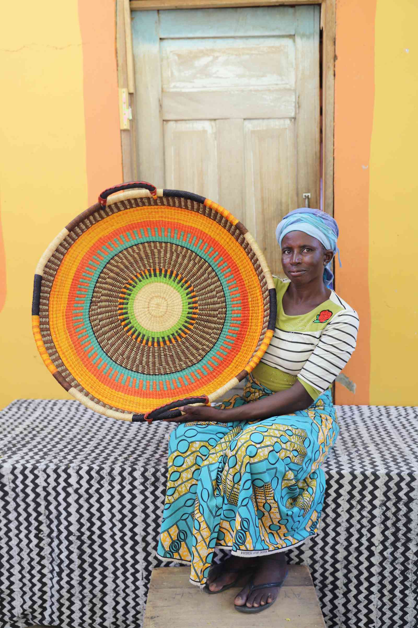 Large handwoven Bolga wall tray disc with orange and teal bands, held by a seated woman in green.