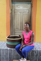 Handwoven Bolga storage basket in dark brown with green stripes, styled beside a seated woman in pink.